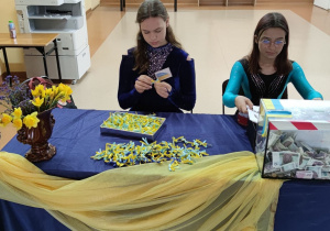 Dwie dziewczynki siedzą przy stole i przygotowują niebiesko-żółte pamiątkowe kokardki.